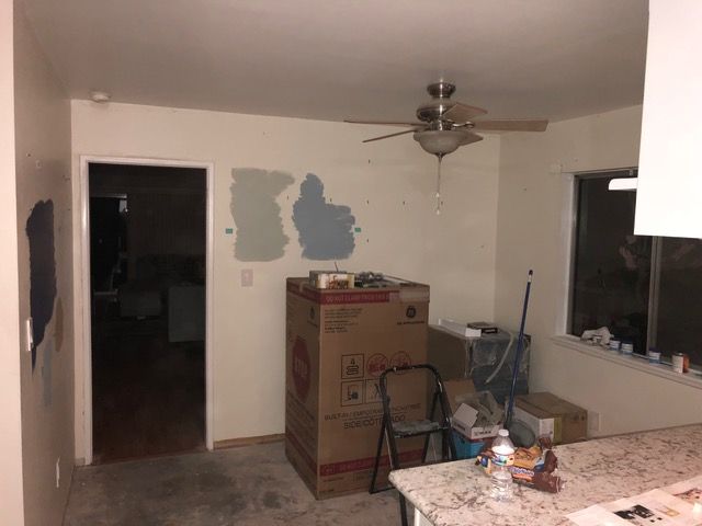 Sparse living room with missing wall paint, cardboard box, ceiling fan, and table by a window