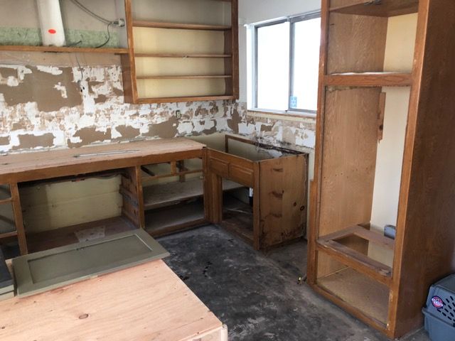 Small unfinished kitchen with worn cabinets, peeling walls, and a window over a bare countertop.