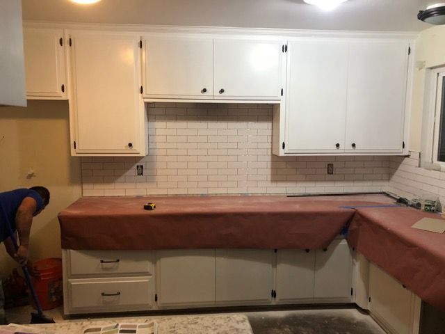 Kitchen under renovation with white cabinets, tiled backsplash, and brown countertop cover; worker at left.