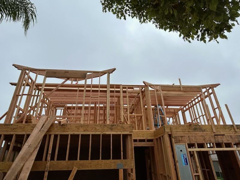 Wood-framed house under construction with exposed framing and roof beams against a cloudy sky
