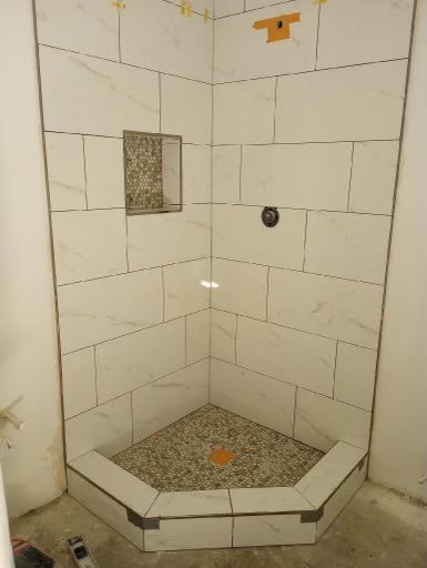 Tiled corner shower with white walls, pebble floor, built-in shelf, and grab bar mount.