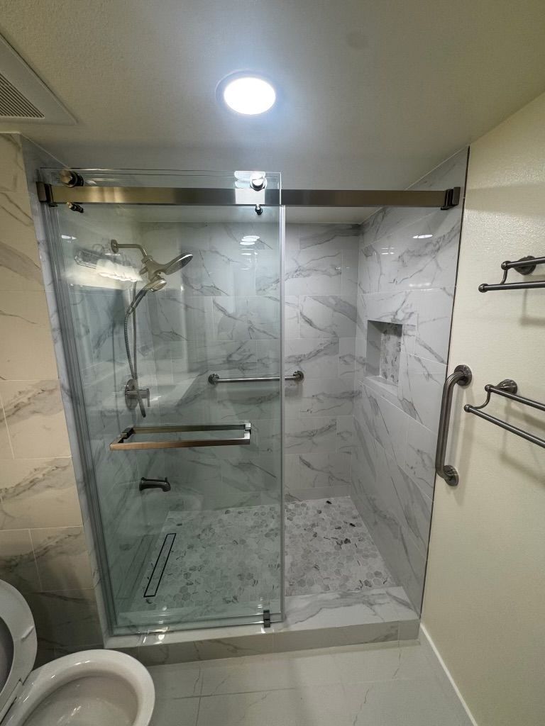 Glass walk-in shower with marble tile, bench, and handheld showerhead in a bathroom.