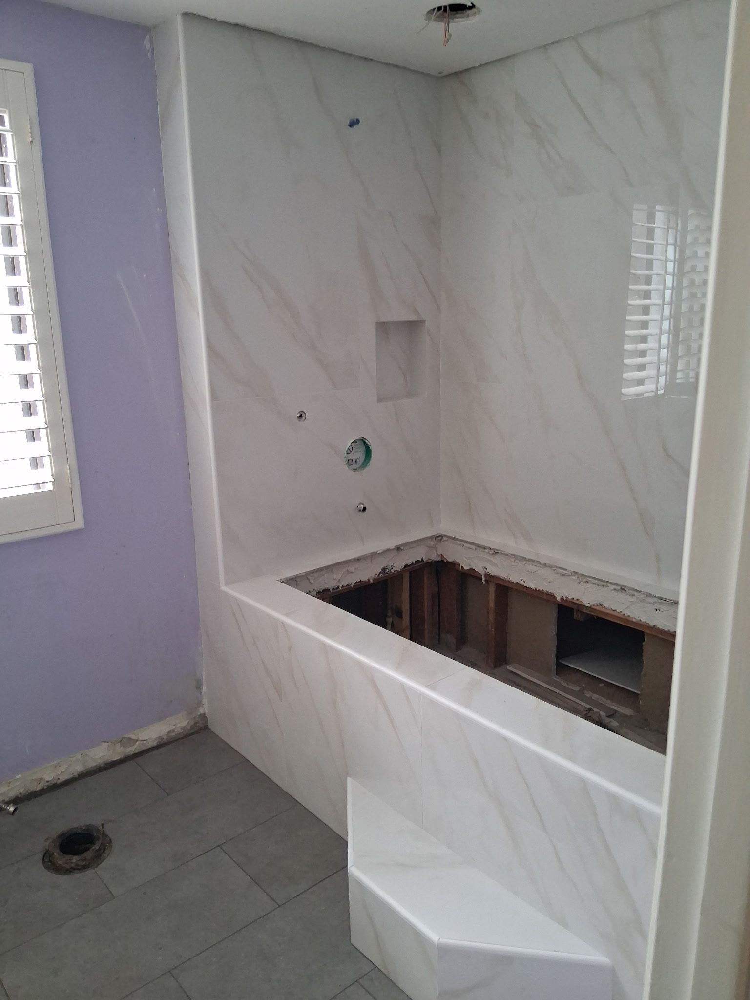 Bathroom under renovation with tiled alcove tub, white marble-look walls, and exposed floor opening