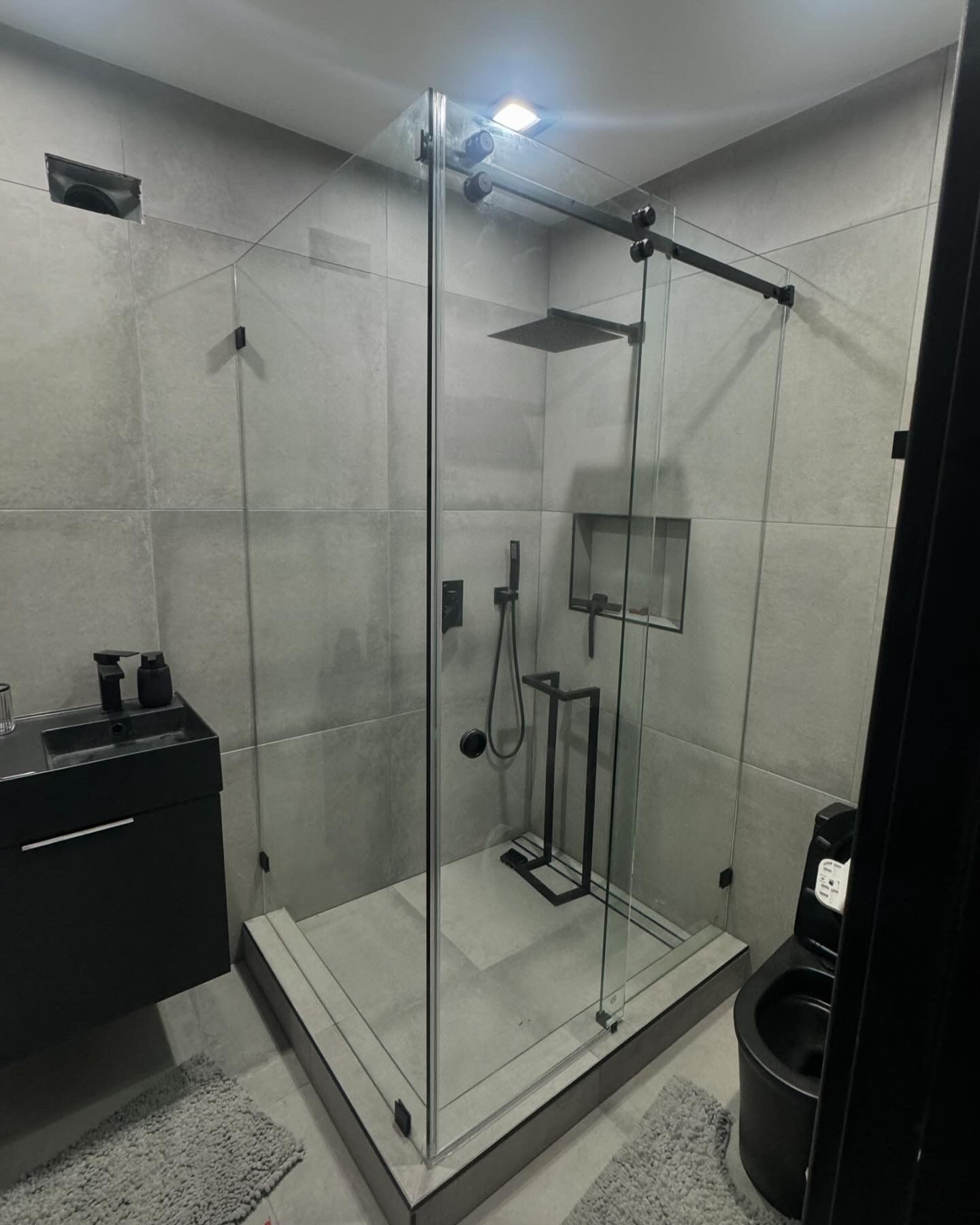 Glass shower stall in a modern bathroom with gray tile, black fixtures, and a rain shower head.