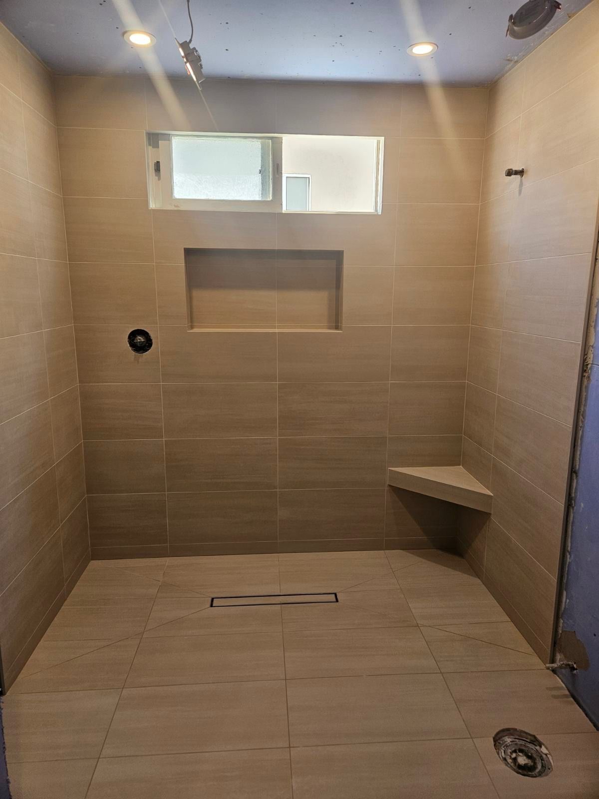 Empty tiled shower with bench, recessed shelf, floor drain, and small high window.