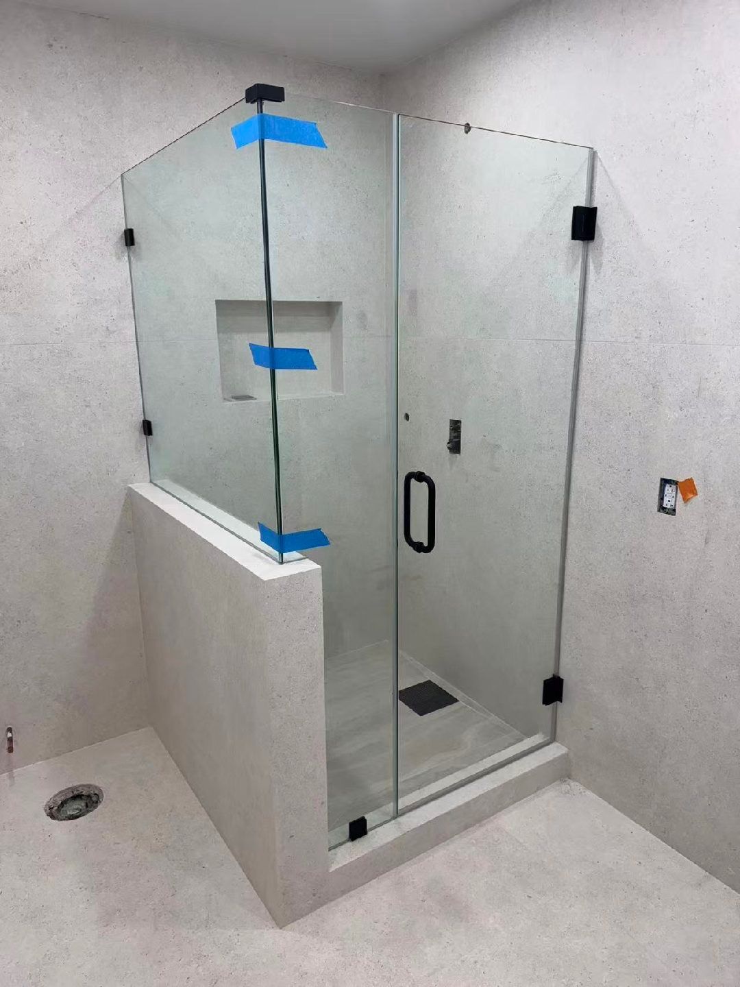 Glass shower enclosure in a tiled bathroom, with a white half wall and black handle, taped for installation.