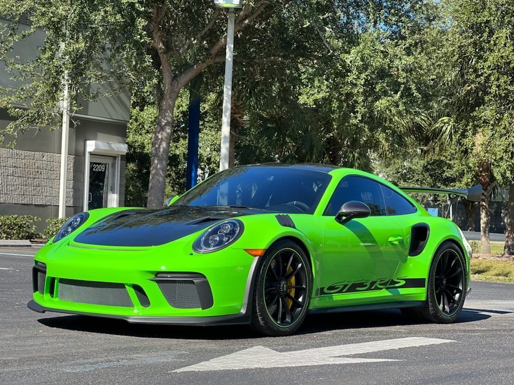 Green Porsche 911 GT3 RS sports car with black accents parked on asphalt. Bright sunlight, trees in background.