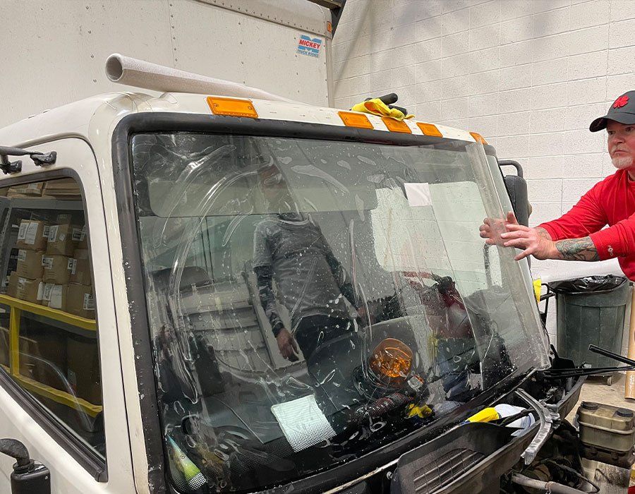 A man in red shirt applies clear film to the windshield of a white truck. Indoor shop setting.