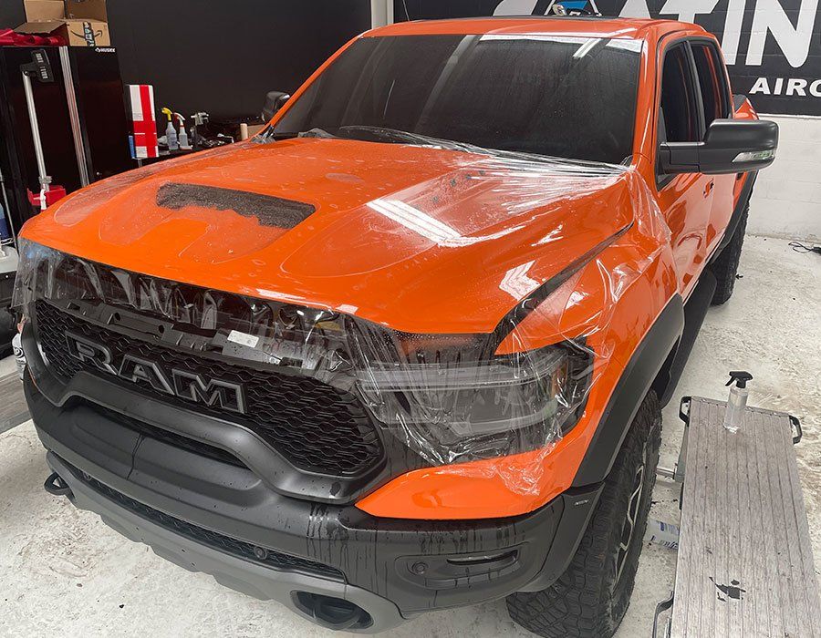 Orange and black Ram truck in a workshop, front view with visible damage on the hood and grille.