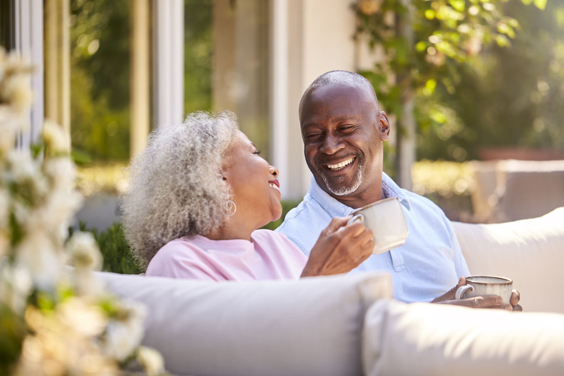 Two smiling adults sit on a couch outdoors, chatting in a sunny garden.