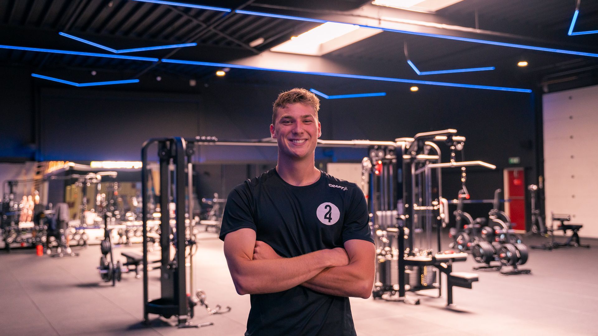 Een glimlachende man, met de armen over elkaar, staat in een sportschool tussen de fitnessapparaten.
