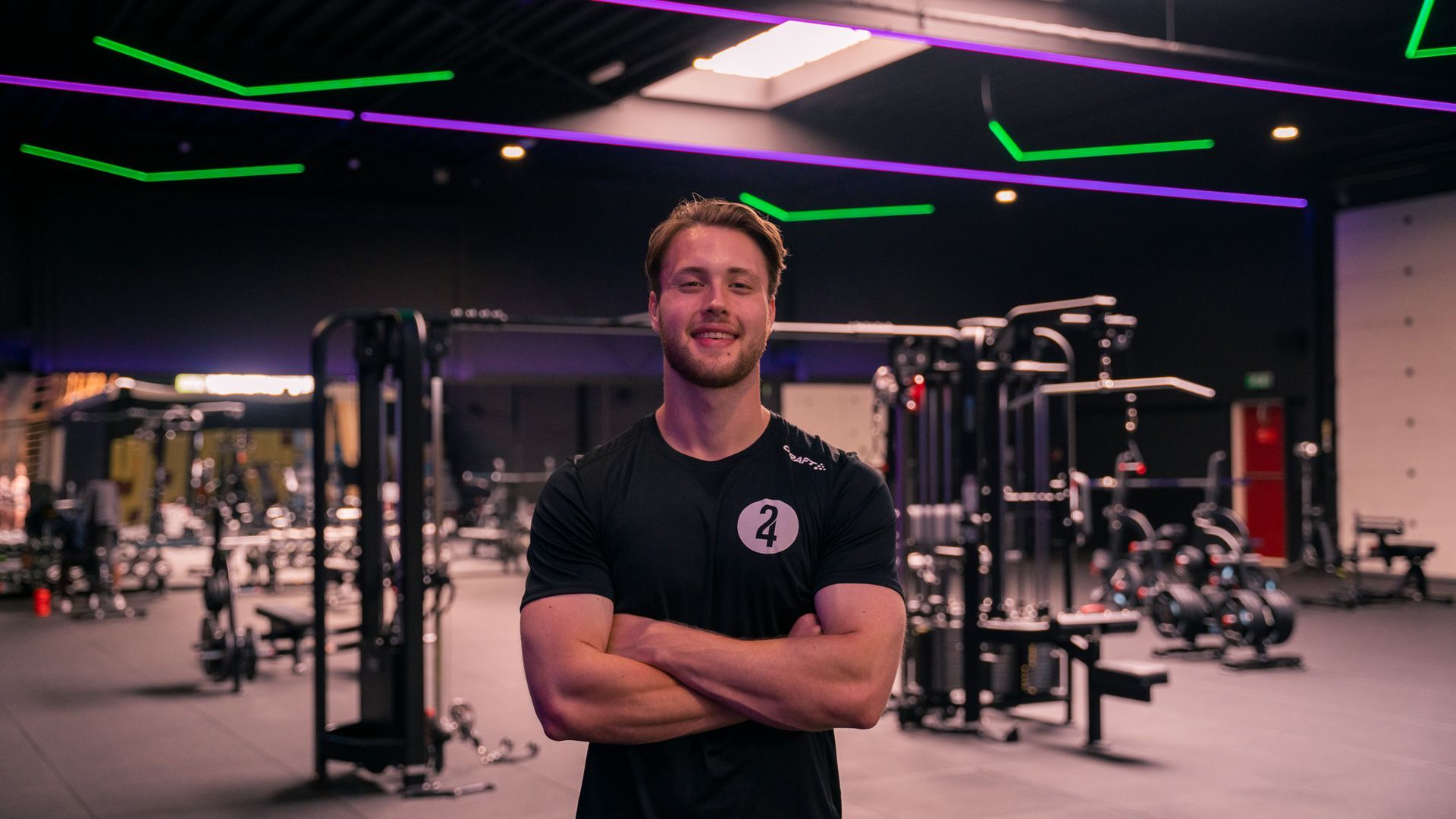 Een man in een zwart shirt met de armen over elkaar, lachend in een moderne sportschool met apparatuur.