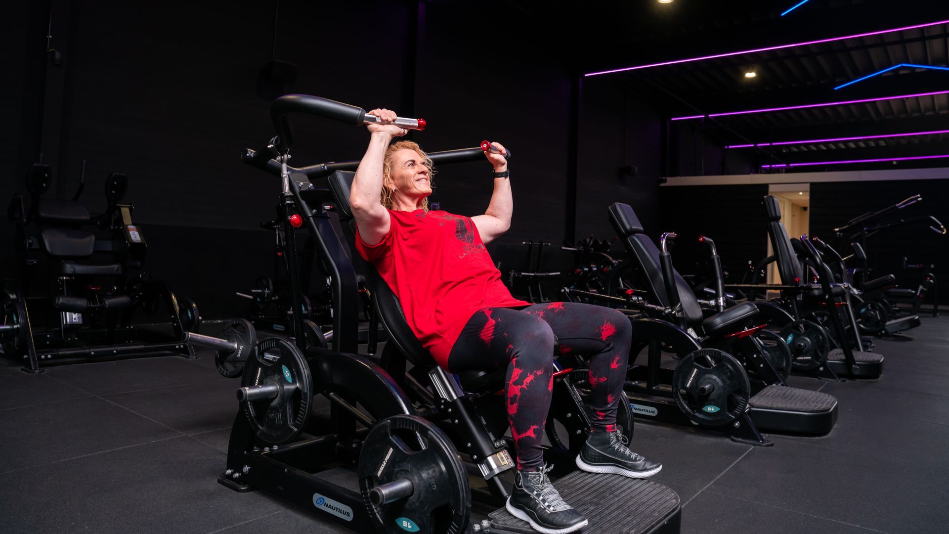 Persoon in rood shirt gebruikt een schouderpersapparaat in zittende positie in een donkere sportschool.