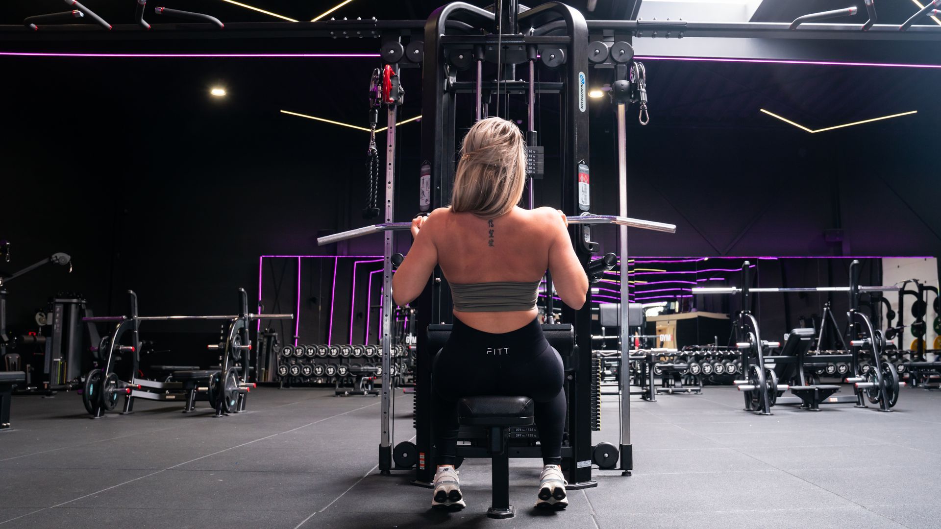 Een vrouw doet lat pulldowns op een apparaat in een sportschool. Ze staat met haar rug naar de camera