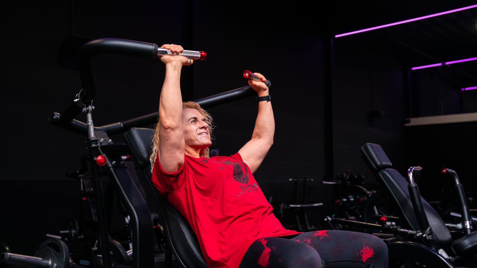 Een persoon zit in een sportschool en gebruikt een overhead press-machine, met de armen omhoog gestrekt.