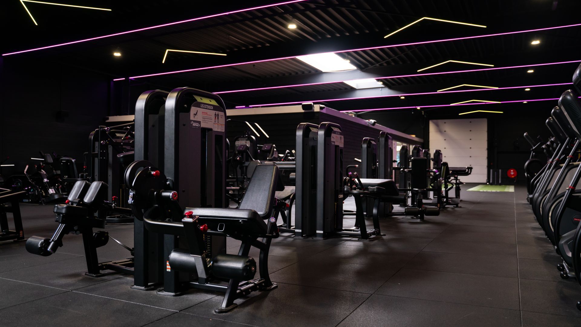 Interieur van de sportschool met zwarte gewichtsapparaten, donkere vloeren en plafond, en roze accentverlichting.