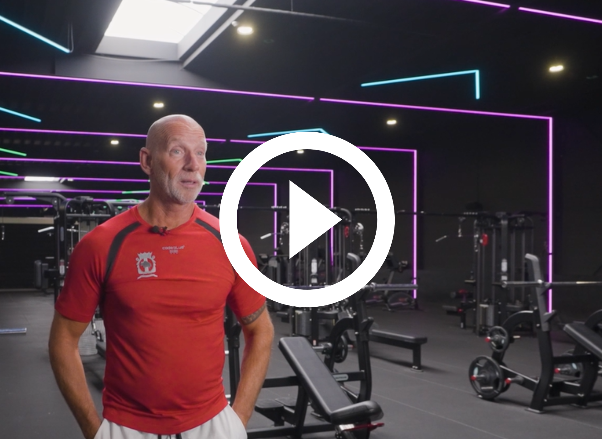 Een man in een rood shirt staat in een sportschool, met fitnessapparatuur op de achtergrond.