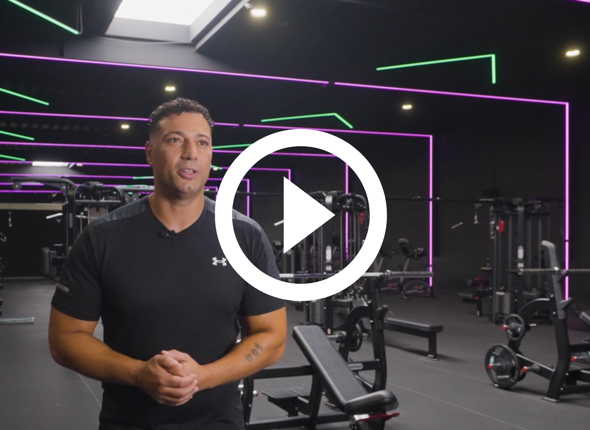 Een man in een sportschool praat; gewichthefapparatuur is zichtbaar, neonverlichting.