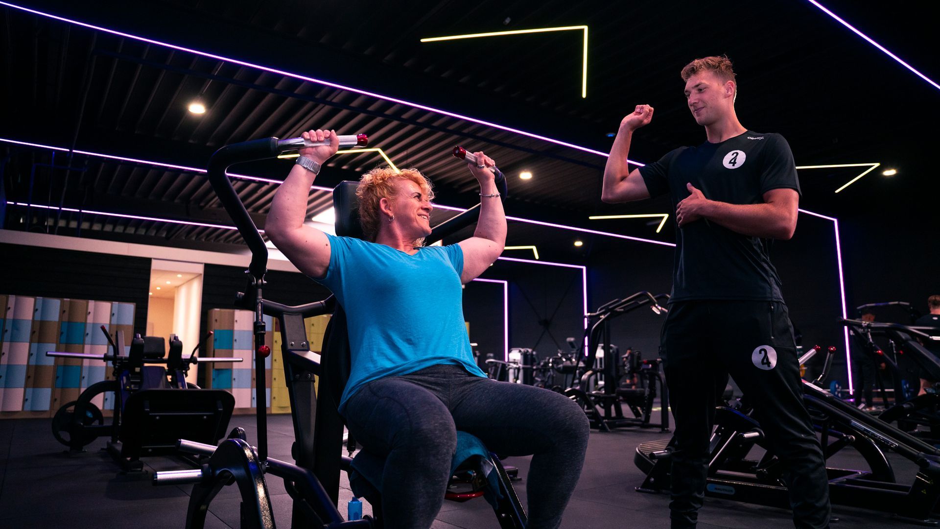 Een vrouw gebruikt een gewichtsapparaat in een sportschool. Een trainer wijst naar zijn biceps.