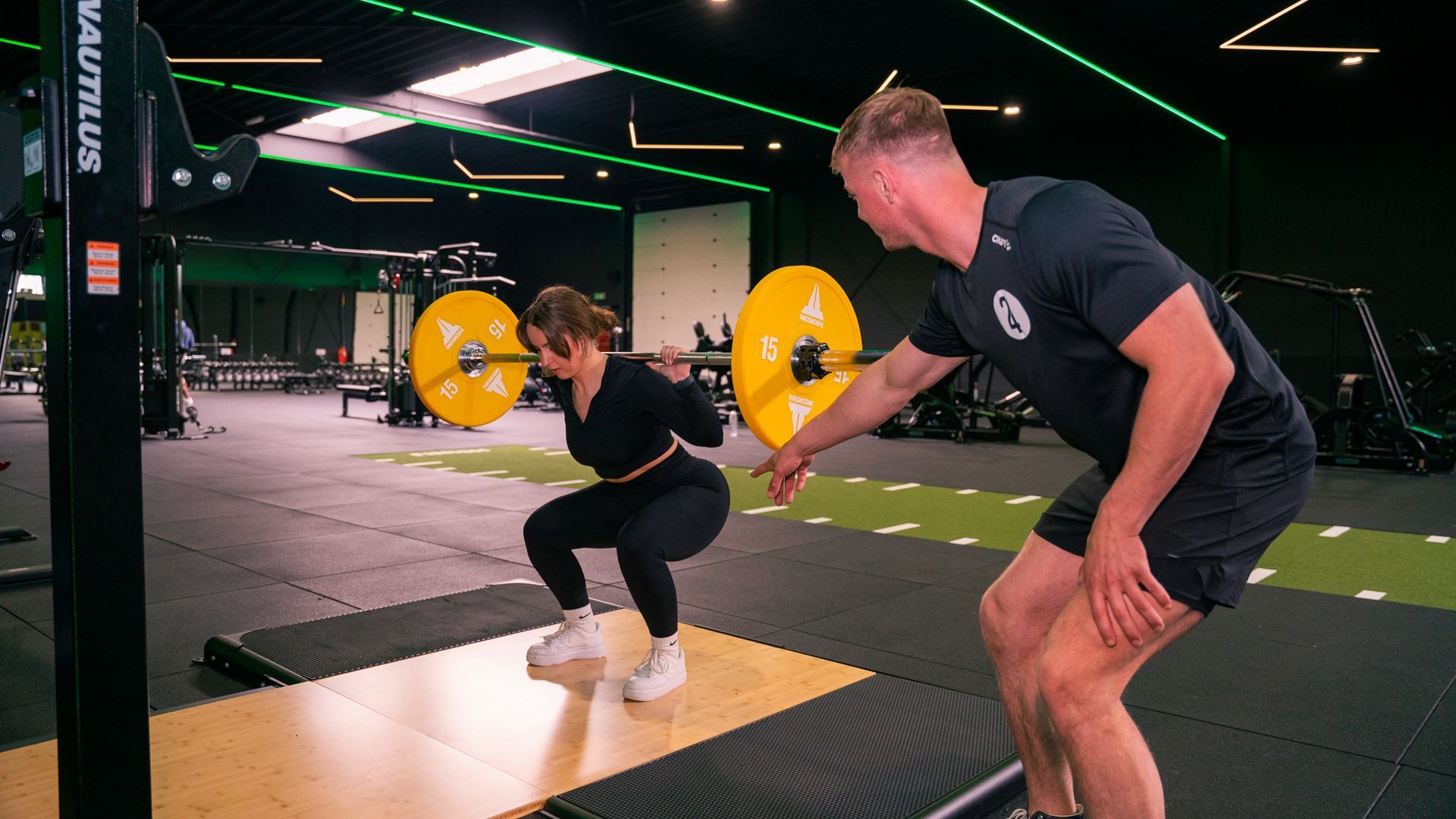 Een vrouw doet een squat met een halter, onder begeleiding van een trainer in een sportschool