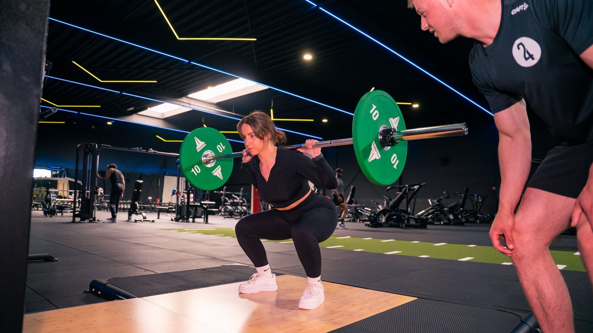 Vrouw doet een squat met een halterstang, terwijl een trainer toekijkt in een sportschool. Groene gewichtsschijven