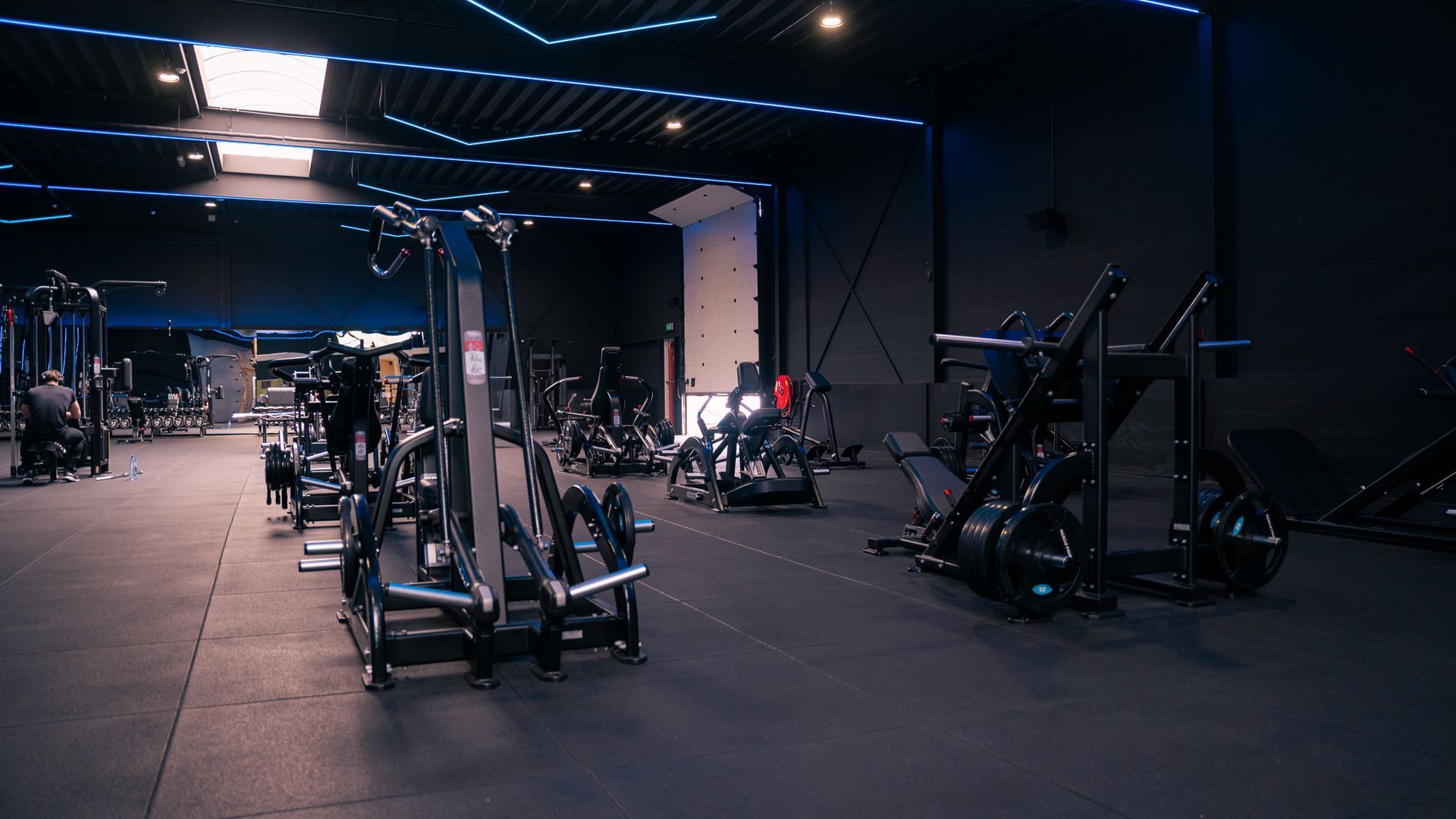 Interieur van een sportschool met diverse fitnessapparaten onder een blauw verlicht plafond.