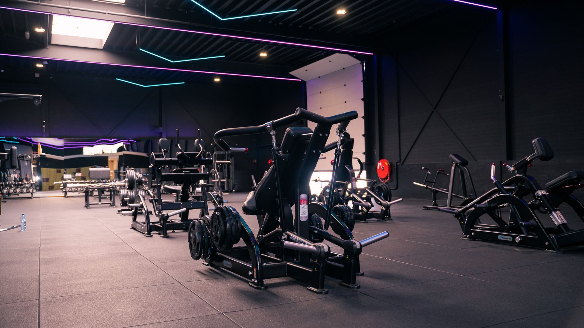 Interieur van een sportschool met rijen fitnessapparaten en een donkere vloer. Paarse en blauwe lampen aan het plafond.