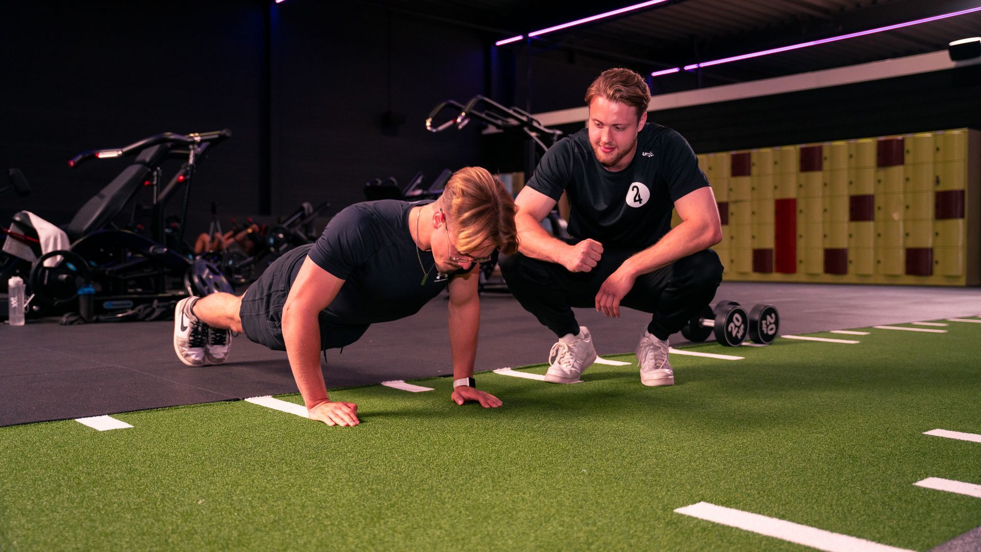 Man doet push-ups op een grasveld met een trainer. Sportschoolomgeving met fitnessapparatuur.