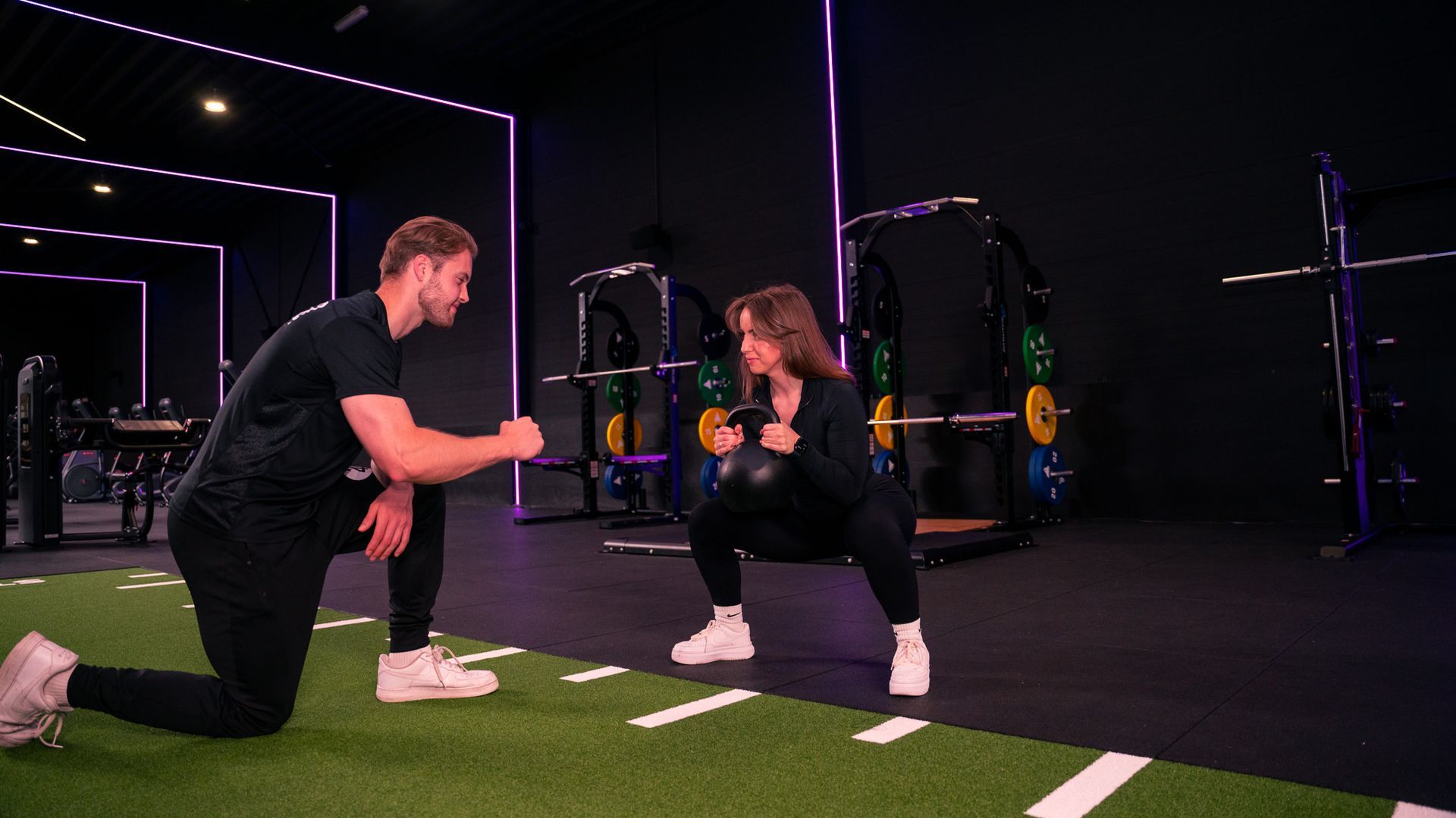 Een trainer helpt iemand met een squat met gewichten in een sportschool.