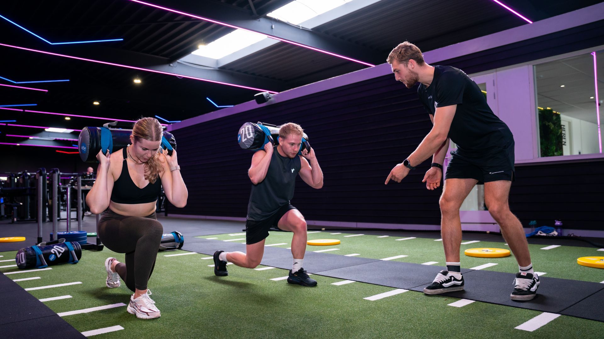 Twee vrouwen en een trainer doen lunges met gewichten in een sportschool