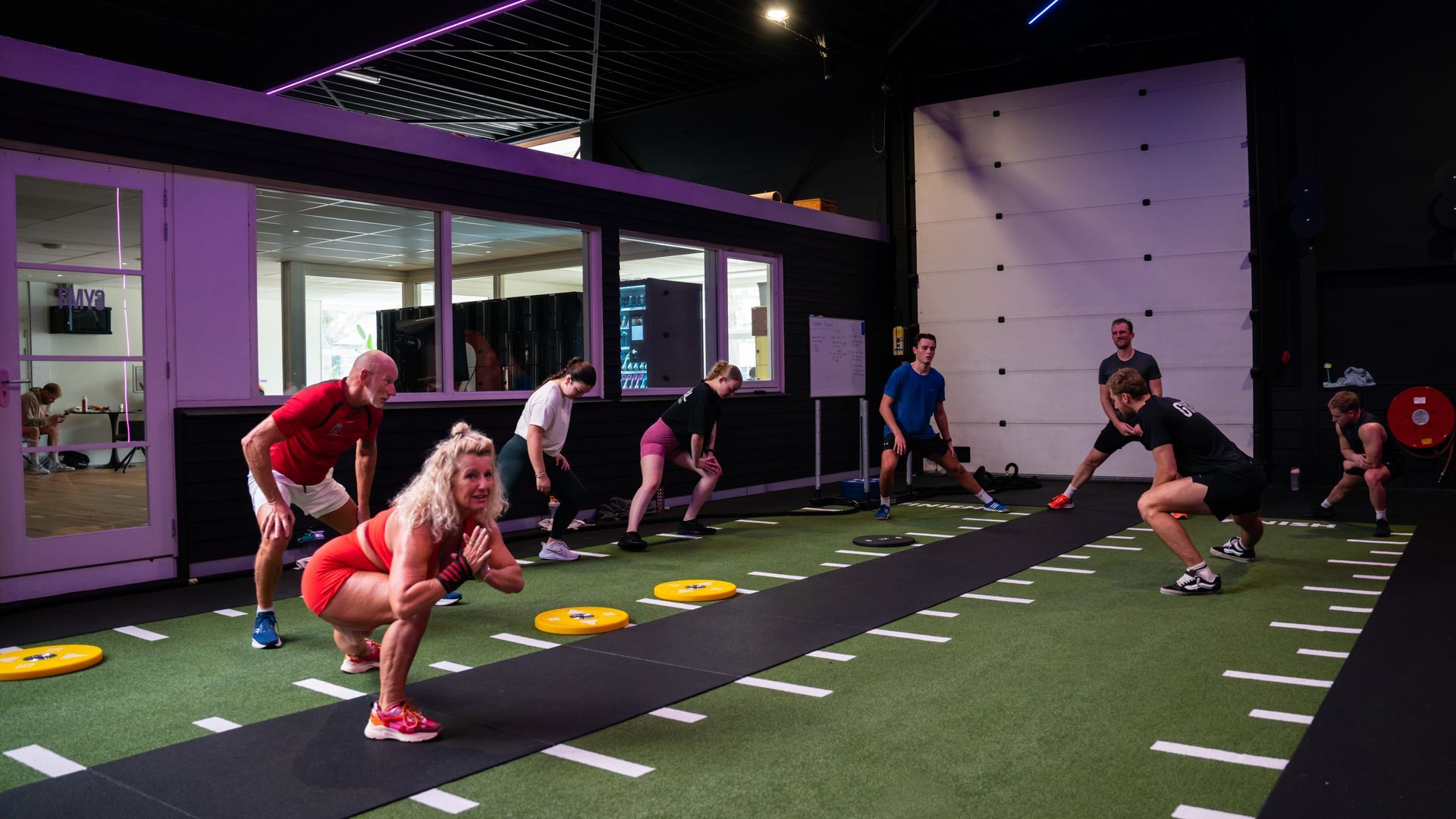 Mensen in een sportschool die oefeningen doen op een kunstgrasbaan. Sommigen doen squats, terwijl anderen toekijken.