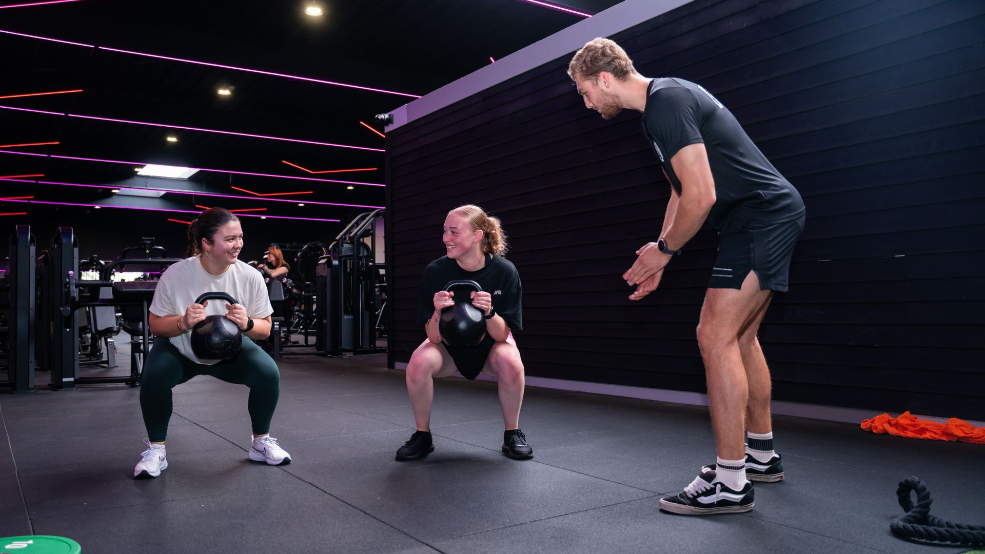 Twee vrouwen doen squats met kettlebells, onder begeleiding van een man in een sportschool.