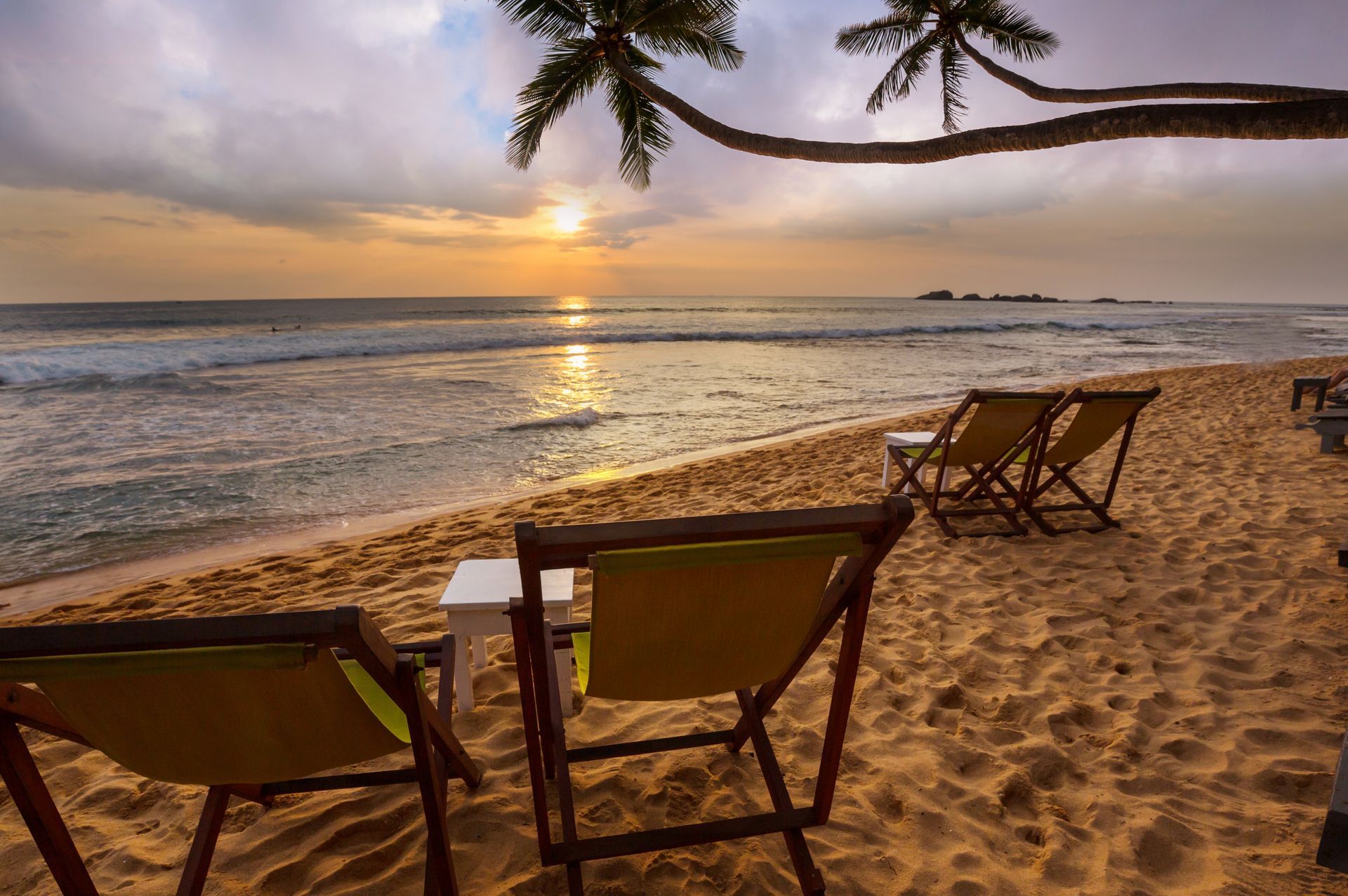 Beach chairs facing a sunset over calm ocean waves, with palm fronds framing the shore