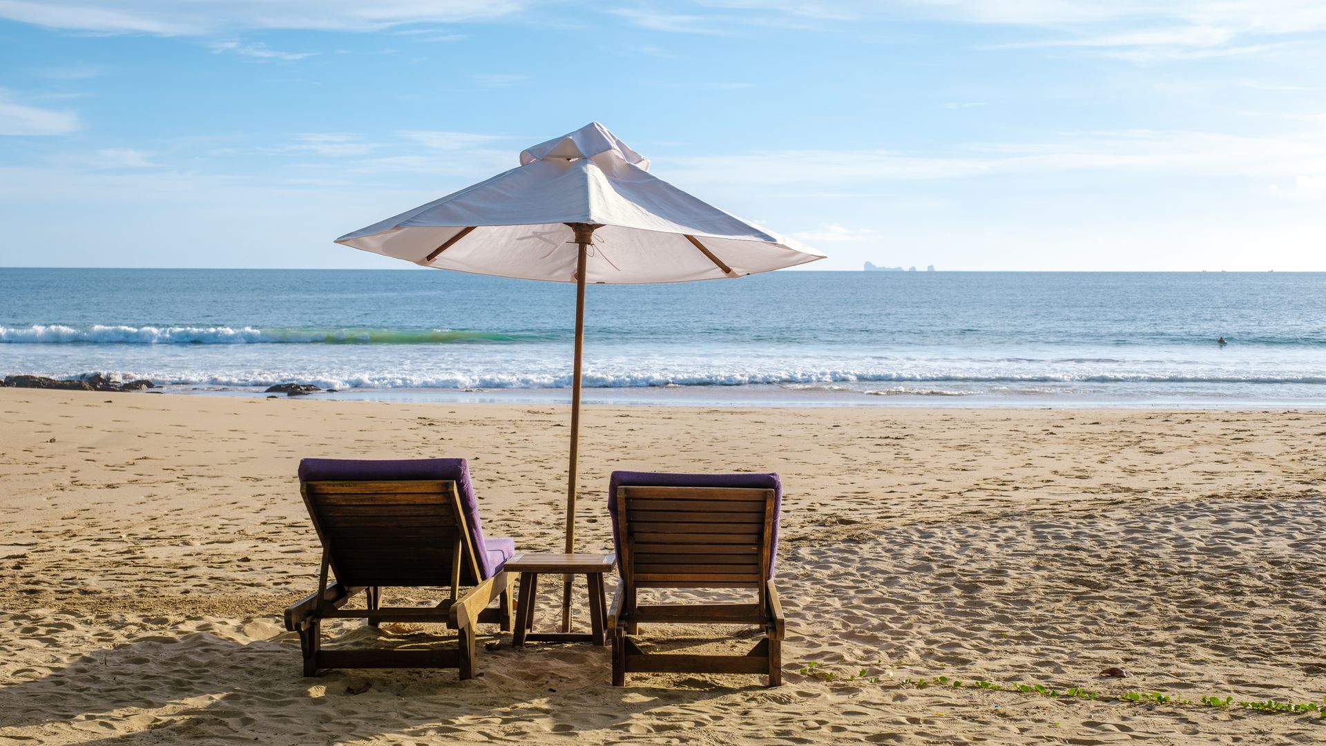 Two wooden lounge chairs under a white umbrella on a sandy beach facing the calm ocean