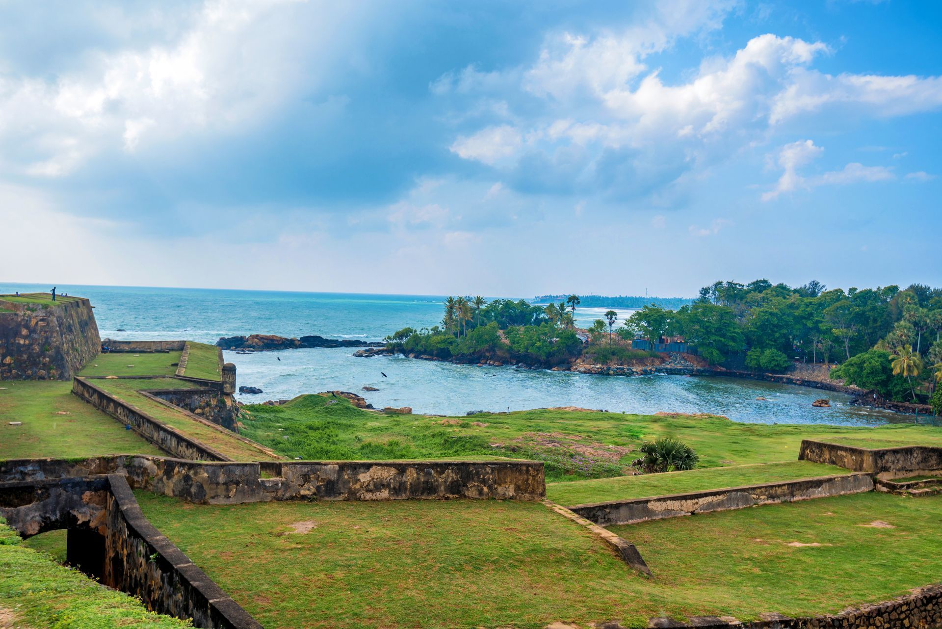 Coastal ruins overlooking a blue sea under a cloudy sky, with green grass and stone walls in the foreground