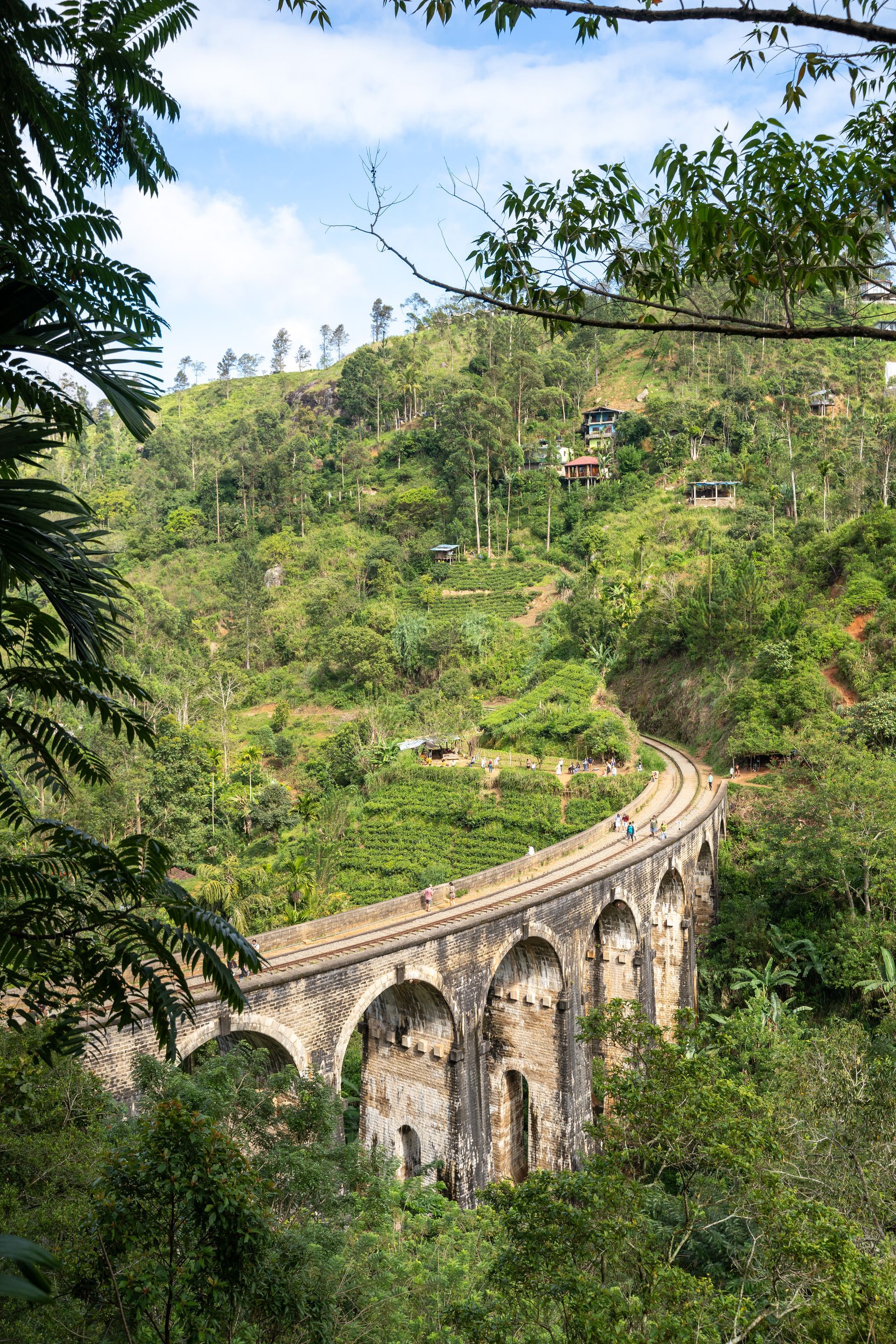 Stone viaduct curving through lush green hillside, framed by trees under a blue sky