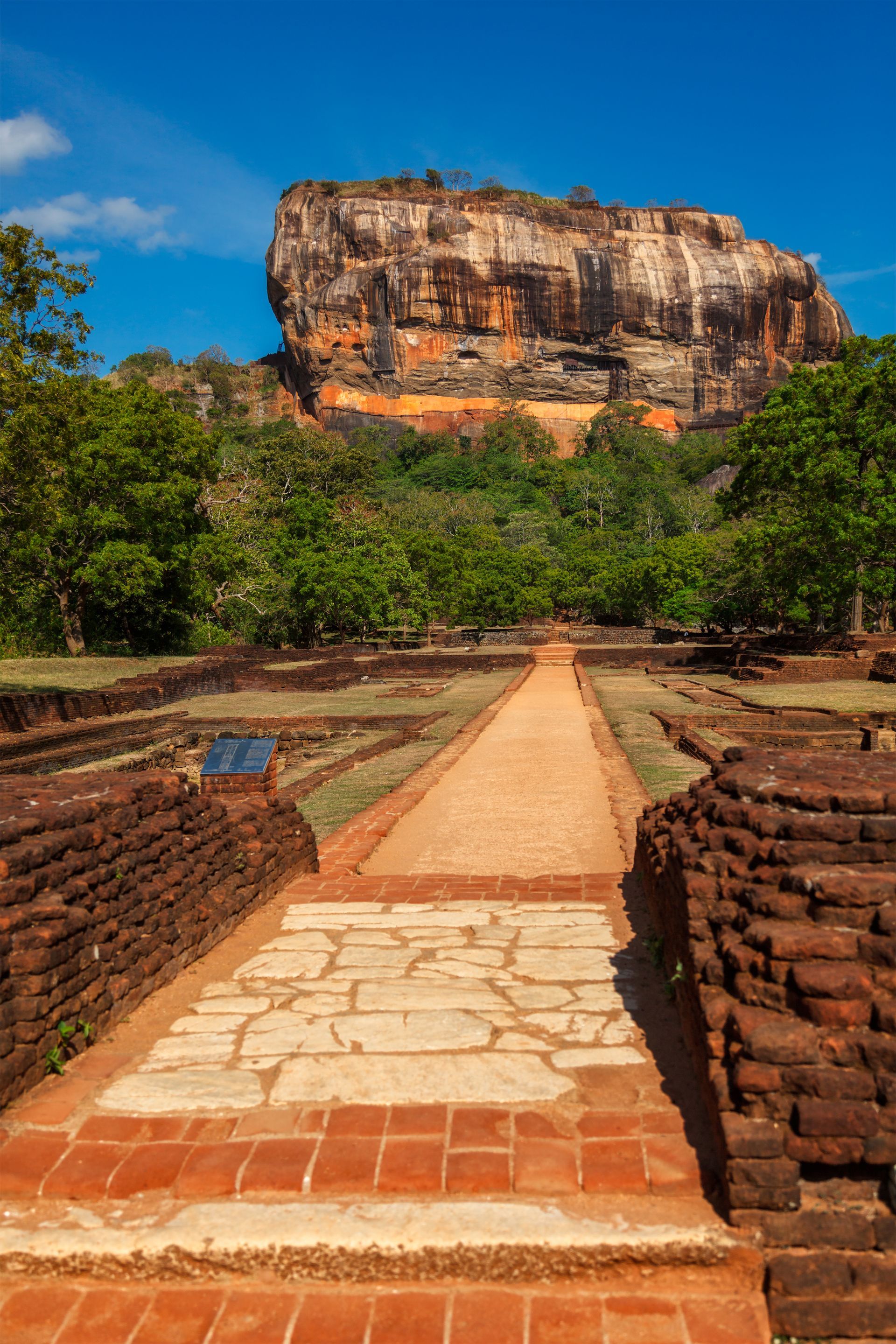 Stone walkway leading to a towering rock cliff under a bright blue sky.