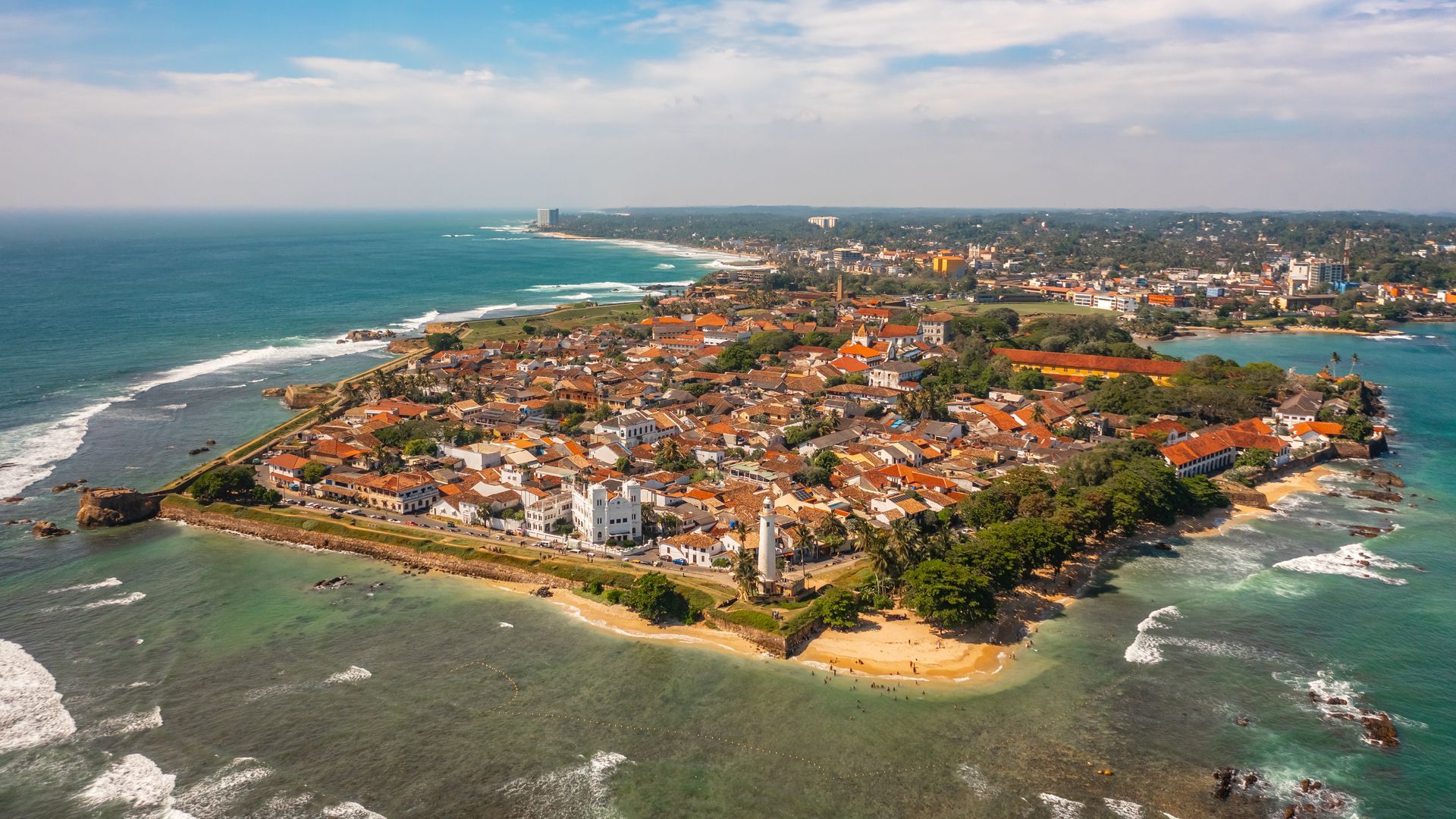 Aerial view of a coastal town on a peninsula with orange roofs, beaches, and blue ocean waves.