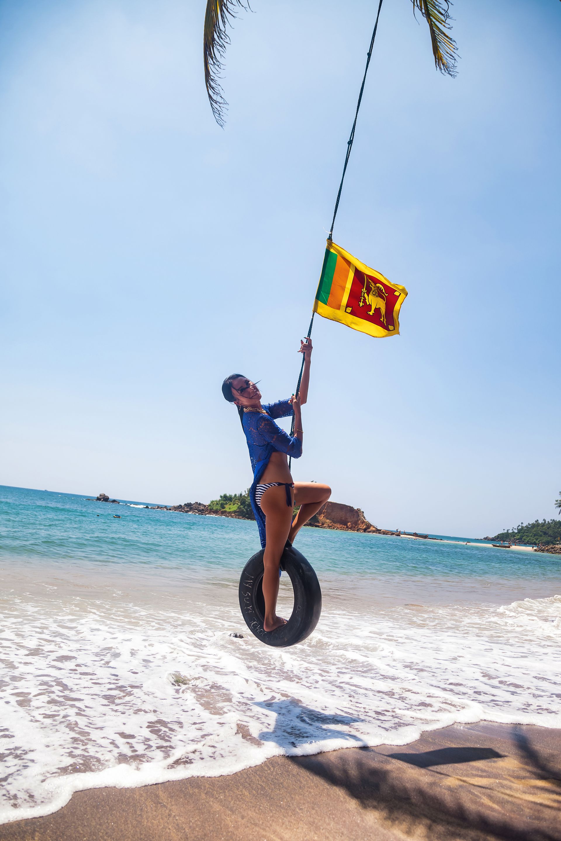 Person swinging on a tire over a sunny beach with turquoise water and a flag overhead