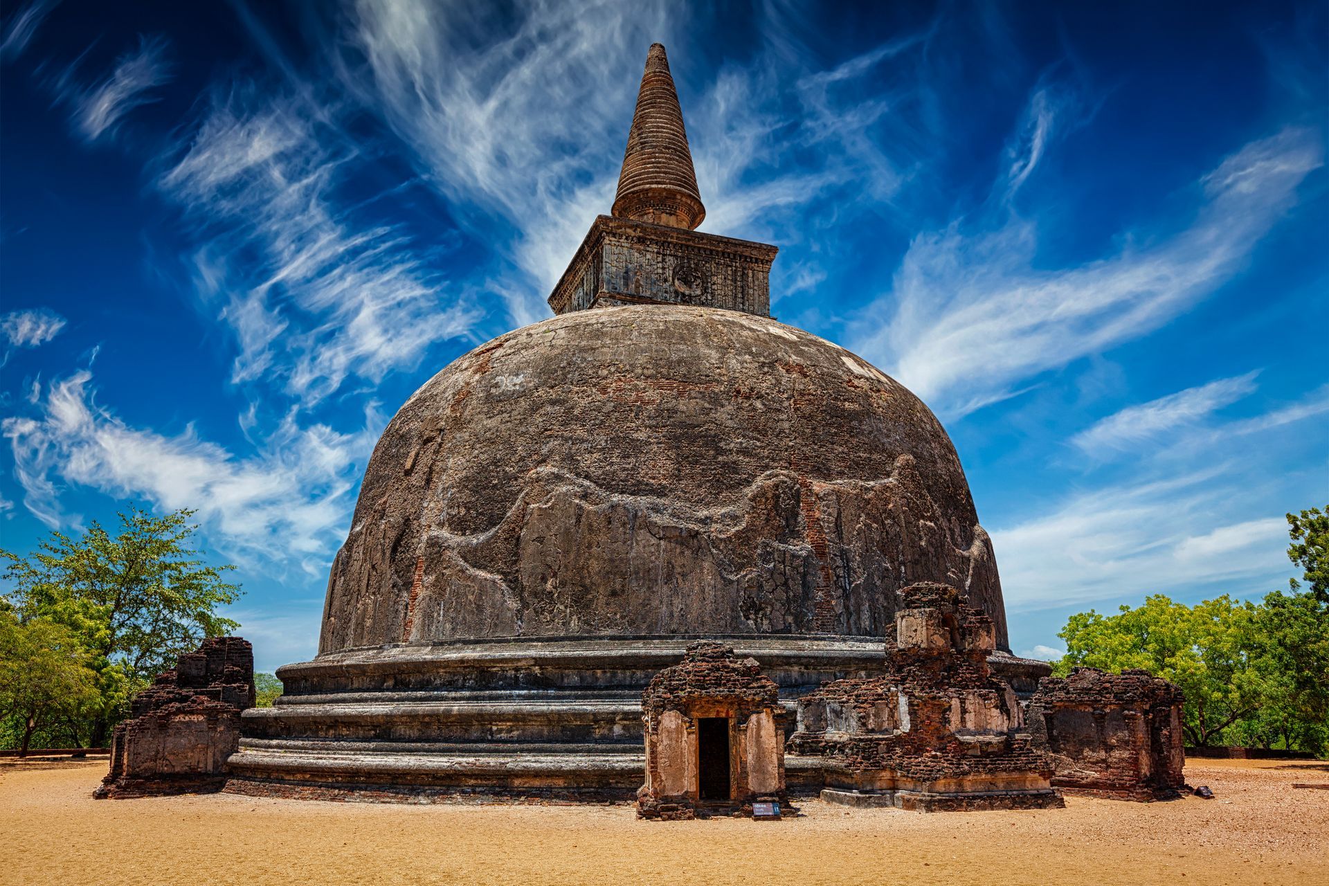 Large ancient stone stupa with a pointed spire against a blue sky, surrounded by steps and trees