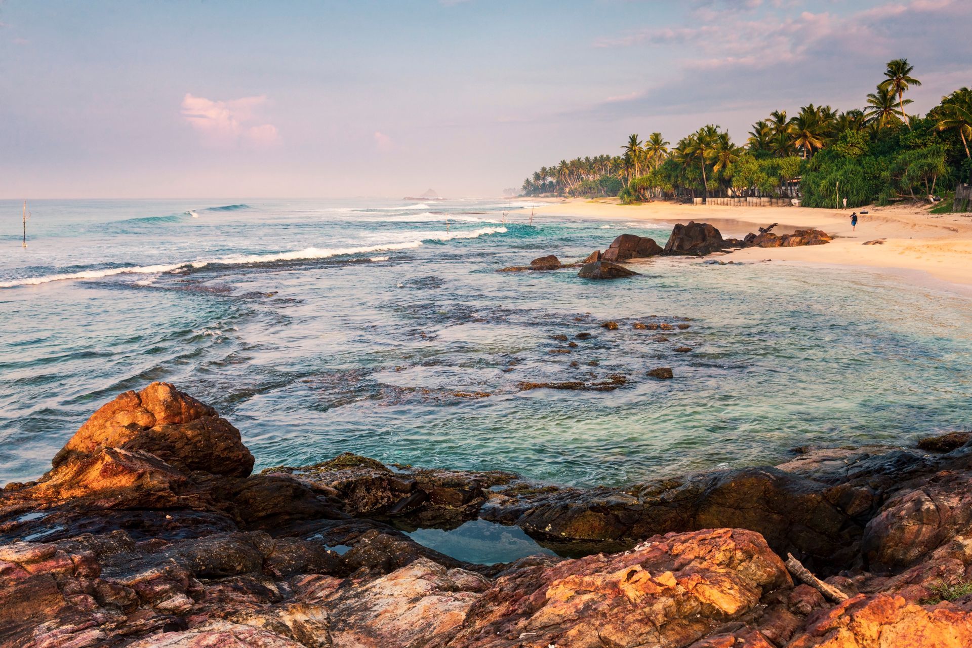 Rocky tropical beach at sunrise with gentle waves, palm-lined shore, and pastel sky