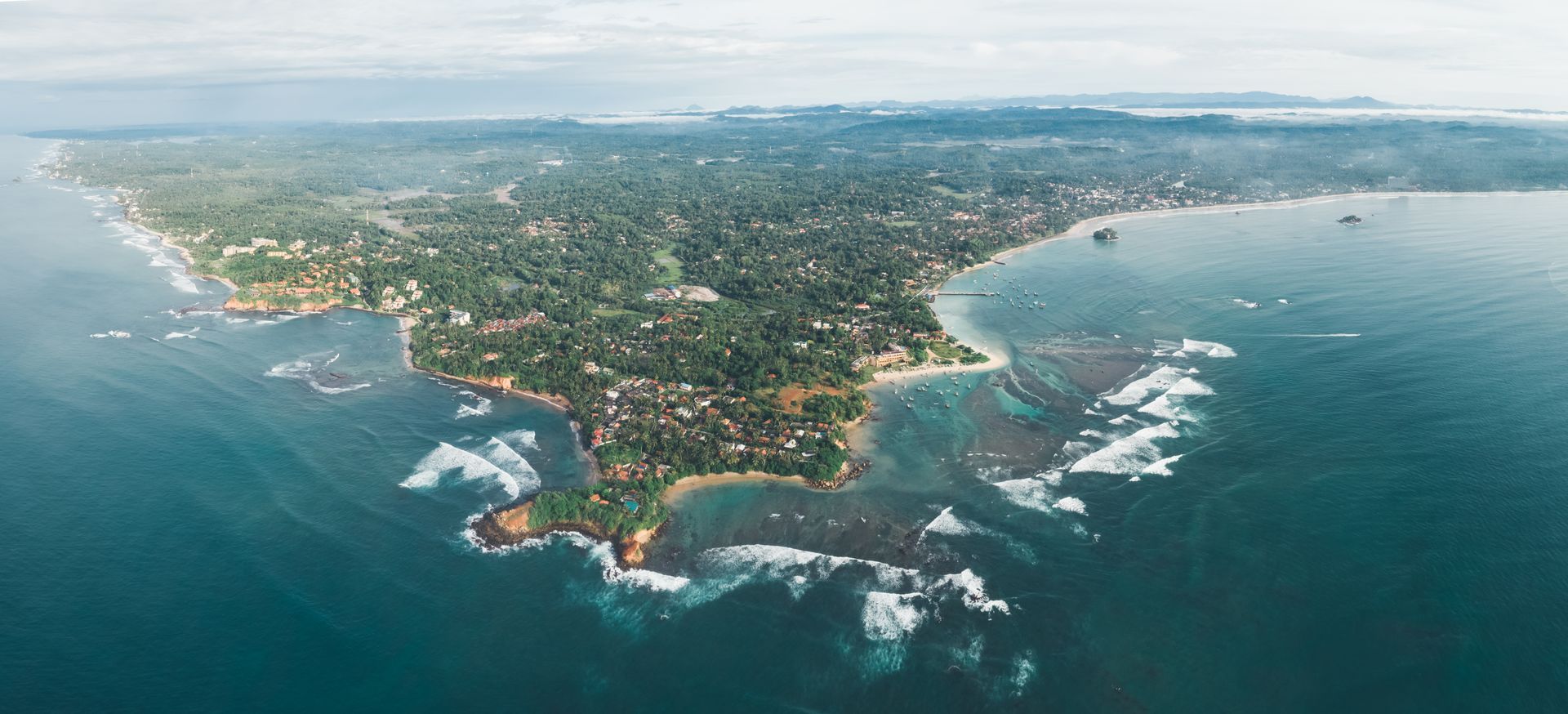 Aerial view of a coastal peninsula with green hills, a town, and waves breaking along rocky shoreline