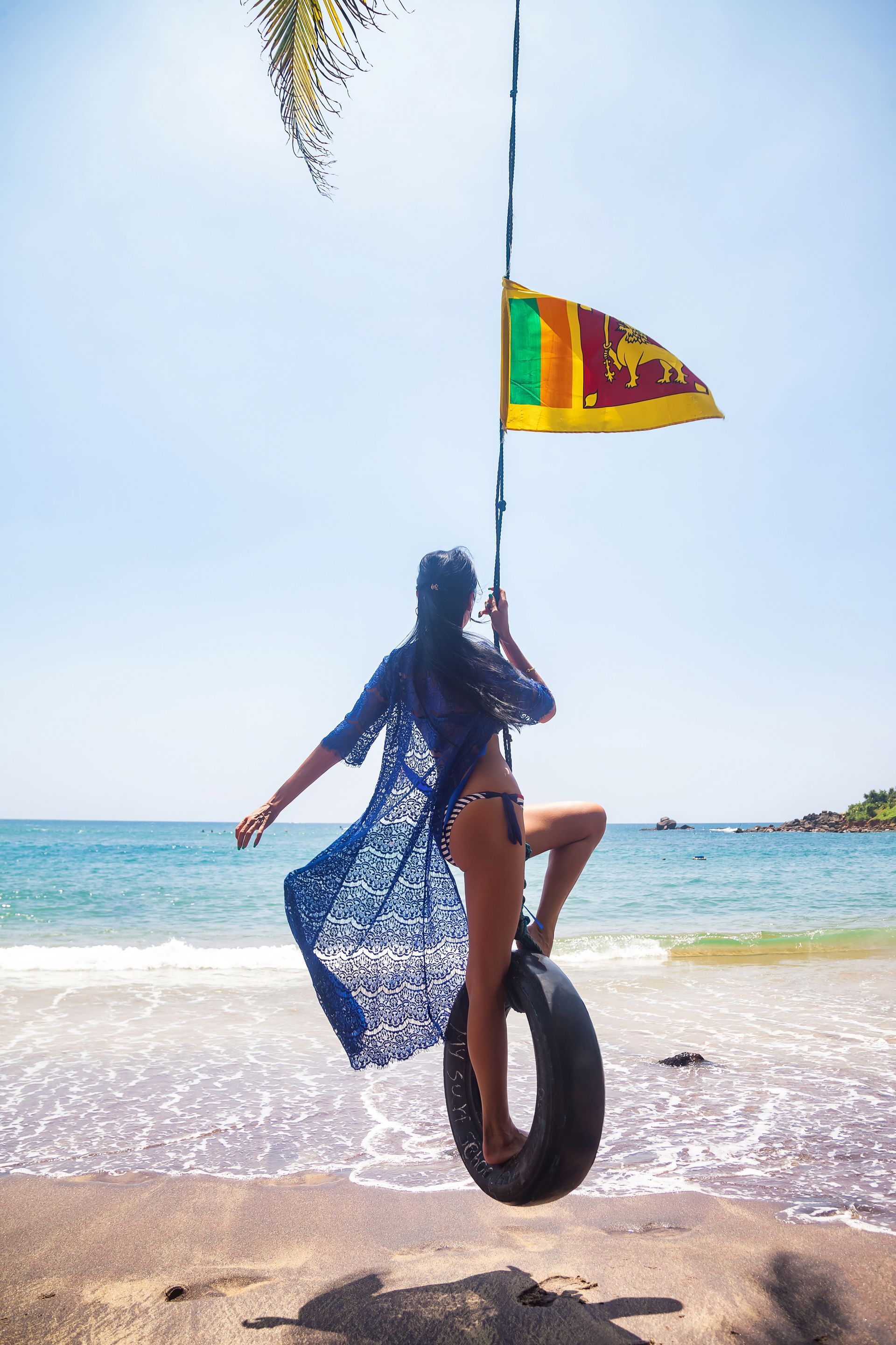 Person on a tire swing at a sunny beach with a colorful flag overhead and ocean in the background