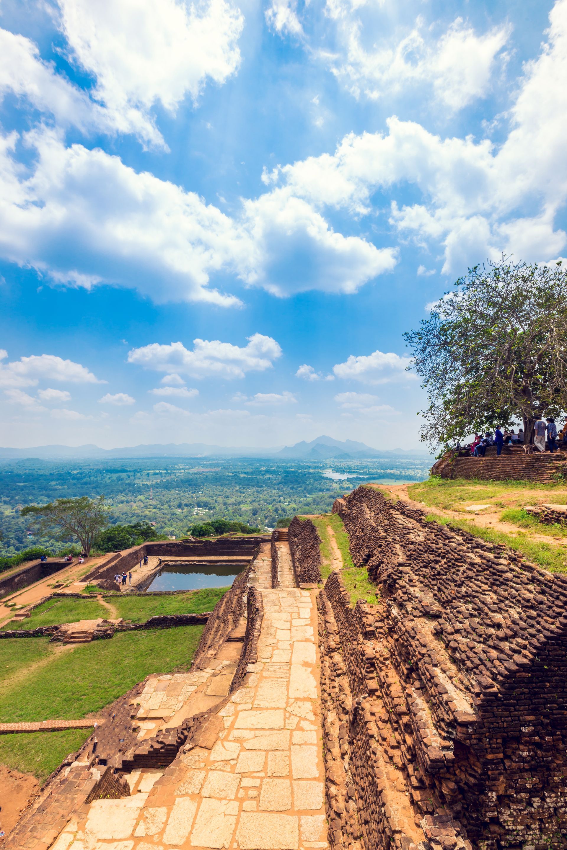 Ancient stone walkway and terraced ruins overlooking a green valley under a bright blue sky with clouds