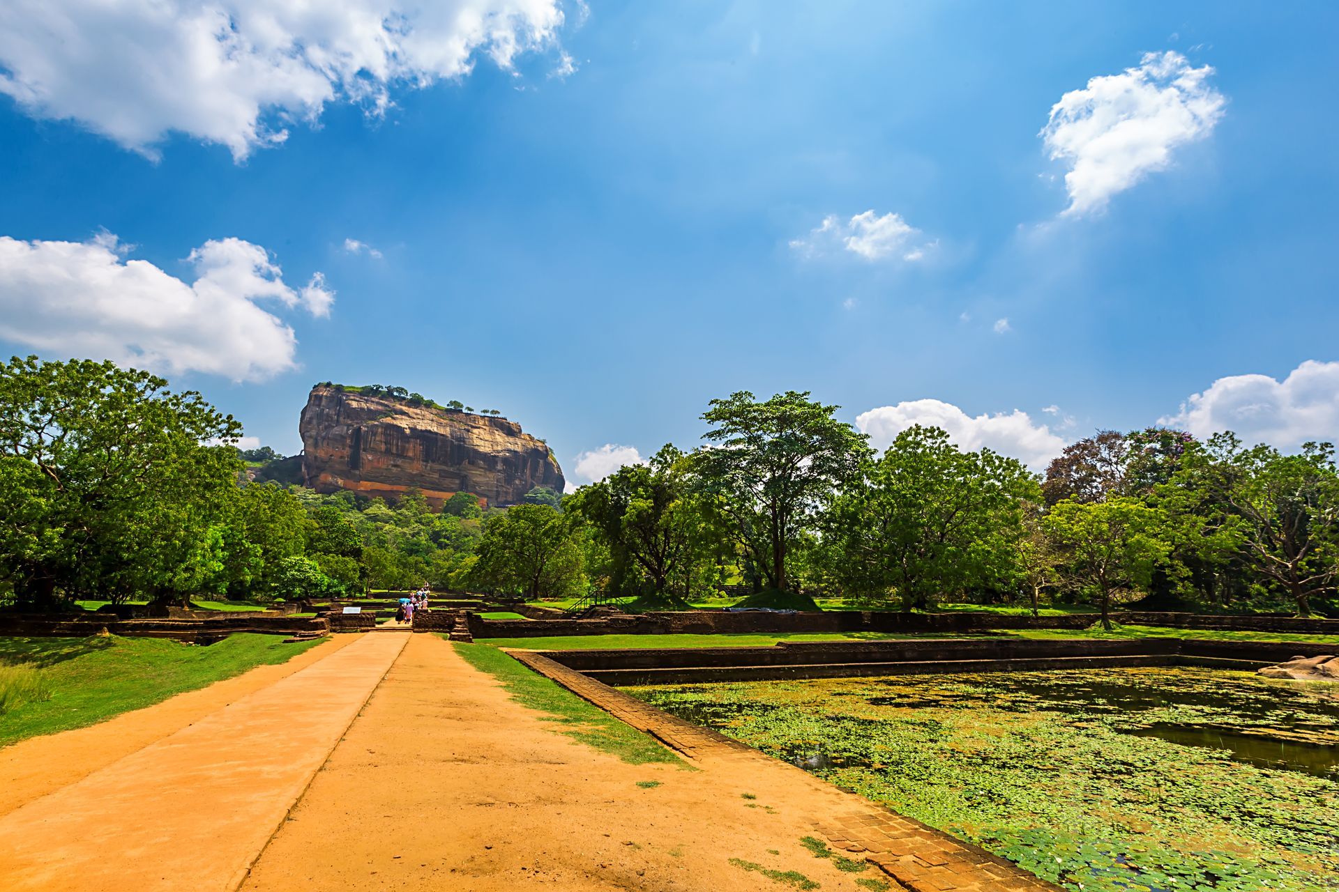 Dirt path through green trees toward a rocky hill under a blue sky with clouds