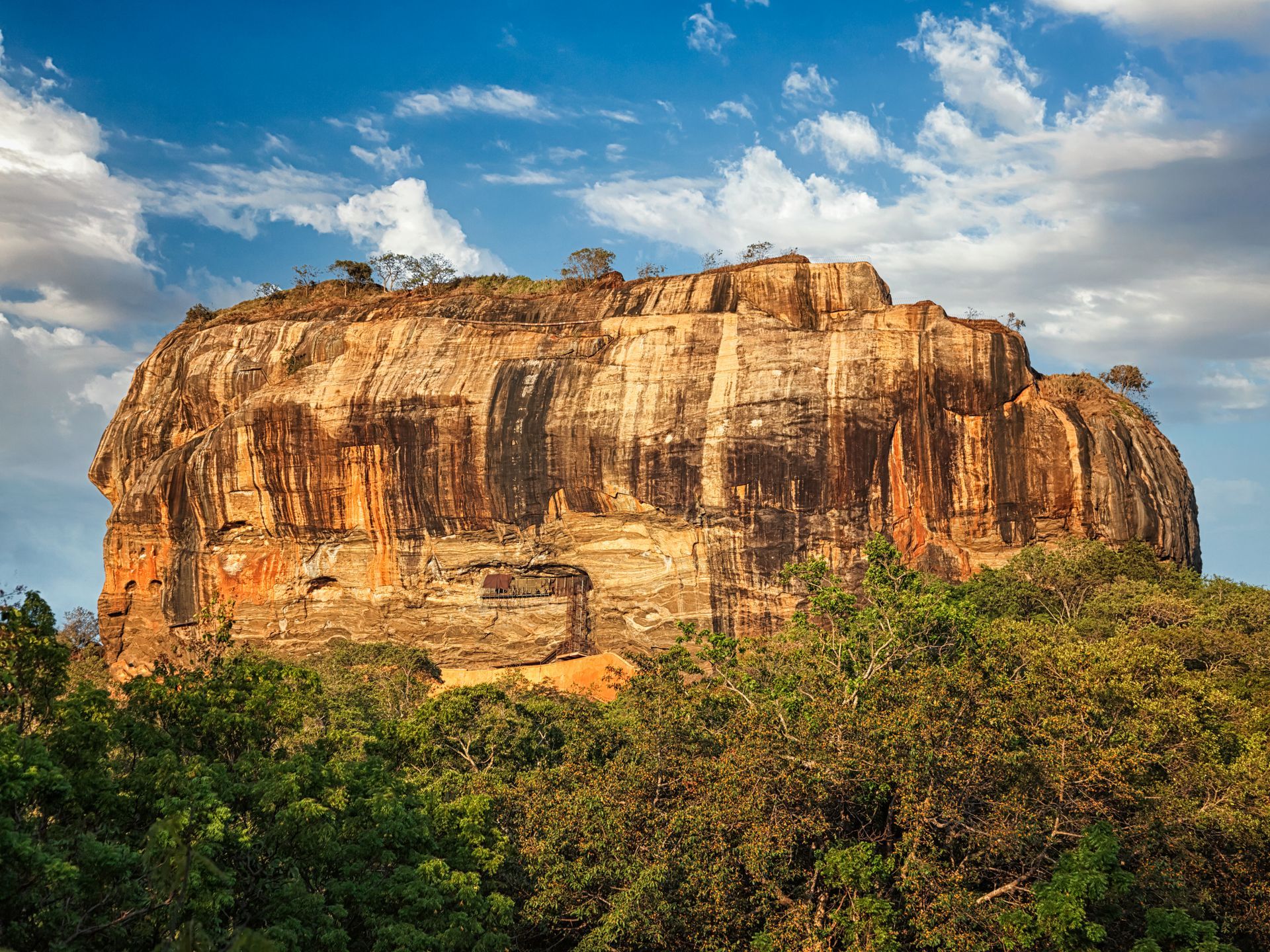 Massive sandstone cliff rising above green forest under a blue, partly cloudy sky