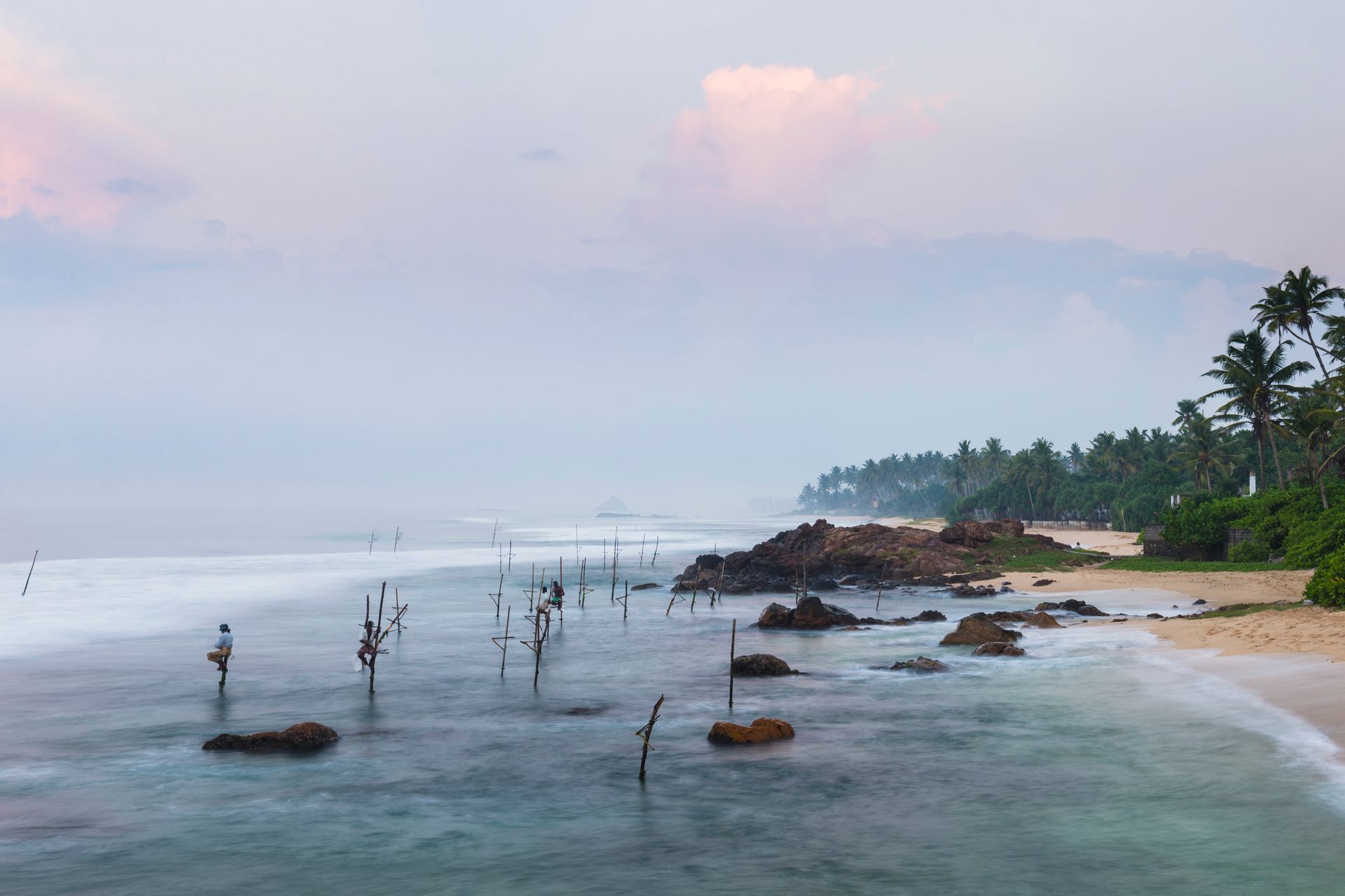 Calm tropical beach at dawn with rocks, wooden stakes, and palm trees along the shore