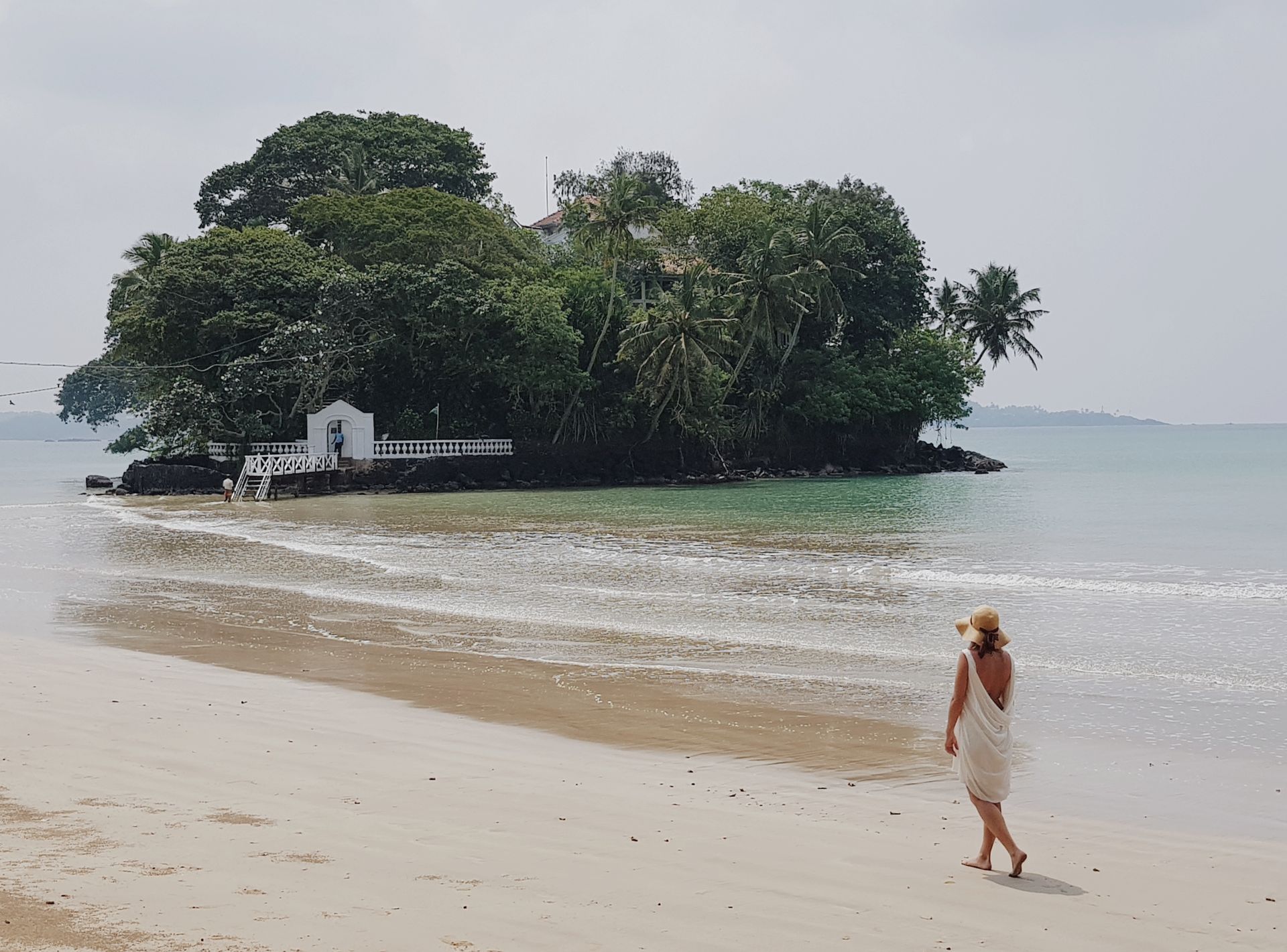 A sandy beach with a small island of dense trees and a person walking near the shore.