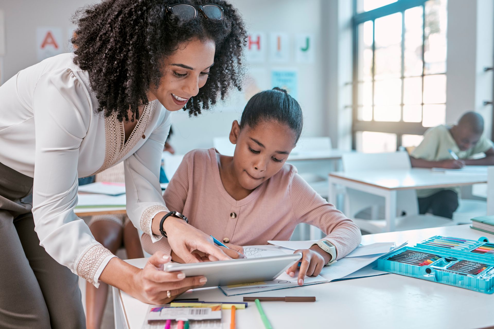 Teacher assisting a student with a tablet in a classroom. Both are smiling.