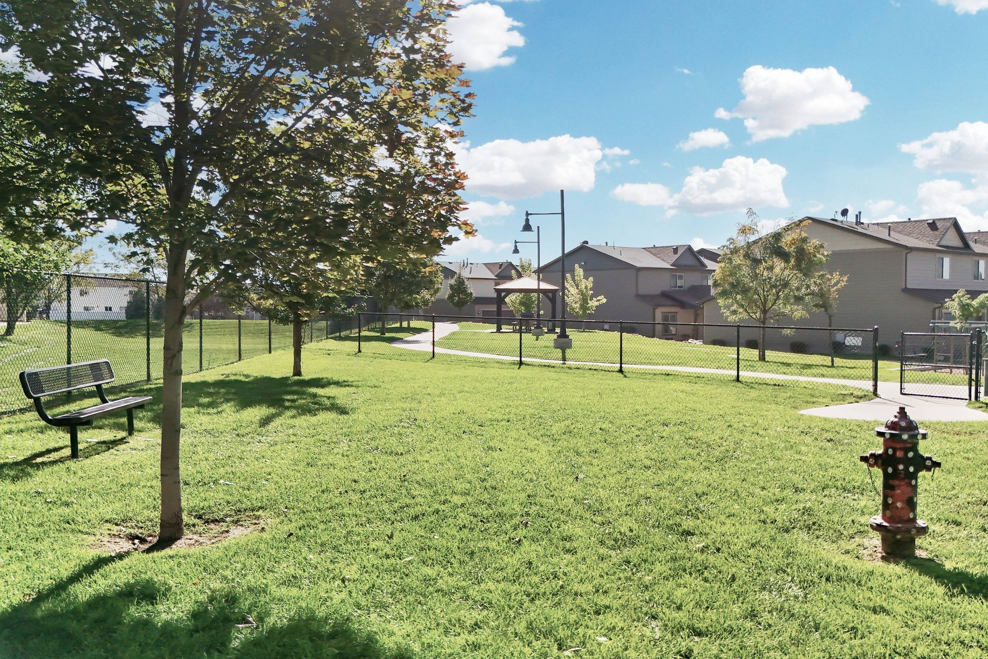 Park with green grass, trees, bench, fire hydrant, and fence, houses in the background, under blue sky with clouds.